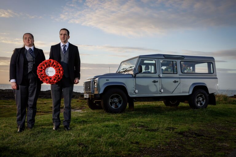 Two funeral directors dressed in formal dark suits stand on grassy terrain holding a large red poppy wreath with a white centre that reads “William Purves.” Beside them is the Land Rover Defender Hearse parked on the grass, with a scenic backdrop of cloudy sky and distant horizon.