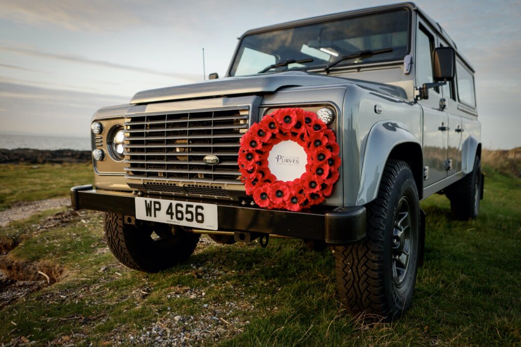 Close-up view of a Land Rover Defender Hearse parked on grassy terrain, with a large red poppy wreath attached to the front grille. The wreath has a white centre displaying the William Purves Funeral Directors logo. The vehicle’s license plate reads “WP 4656,” and the background shows an open landscape under a partly cloudy sky.