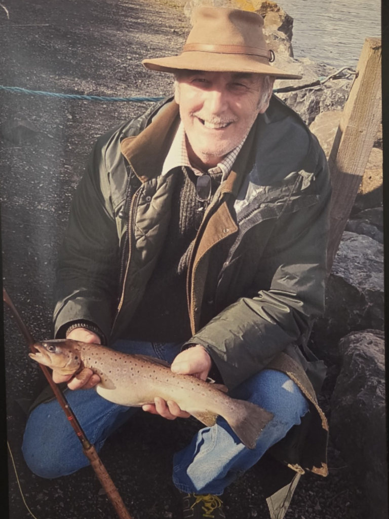 Portrait photograph of a man sitting in fisherman's attire, holding a catch and smiling to the camera. 
