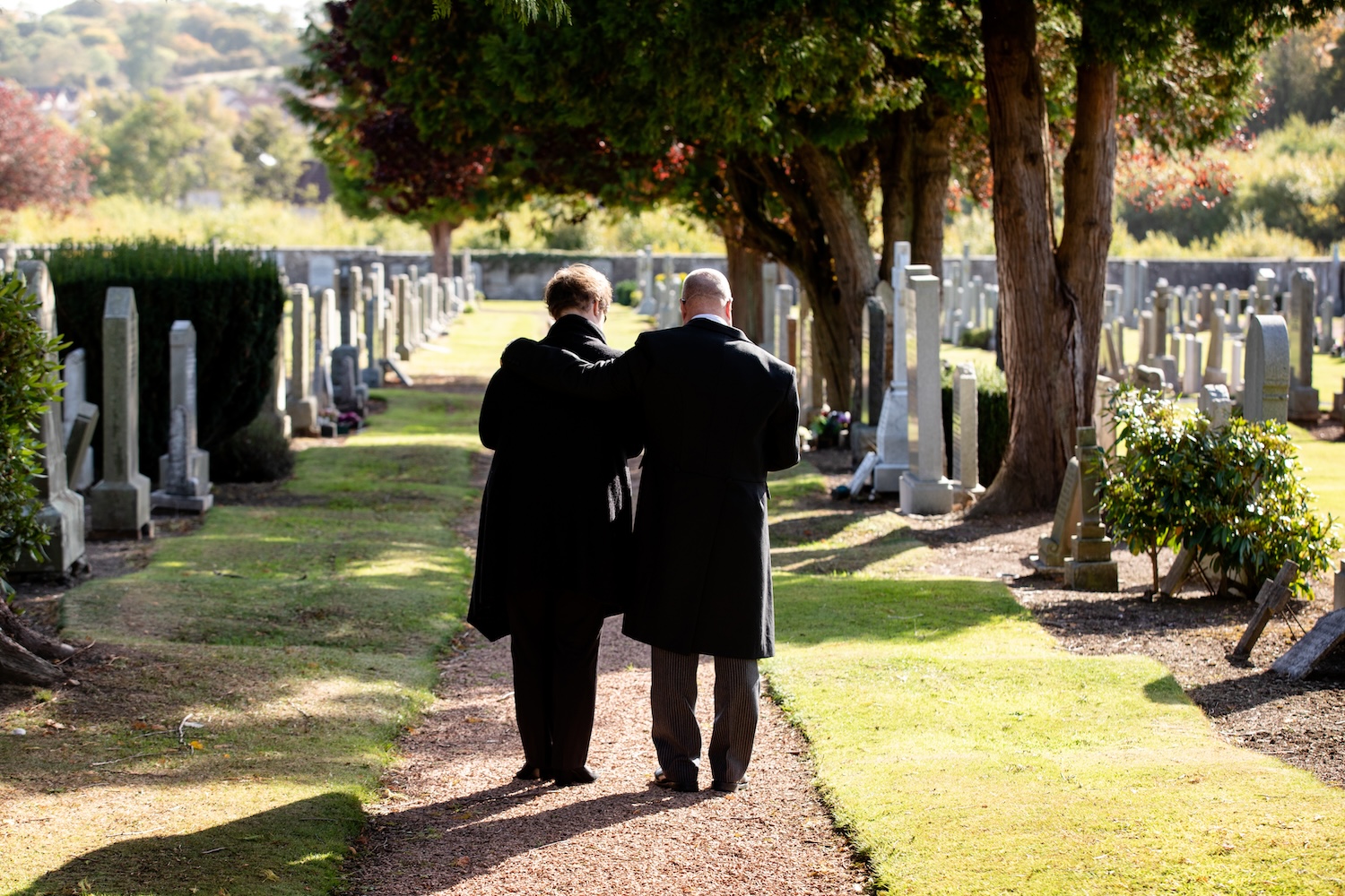 Two people walk along a path in a grave site, with one arm around the other